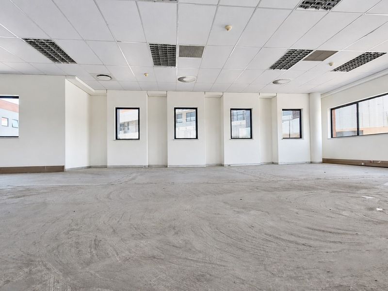 A spacious, open-plan office area featuring a suspended ceiling with recessed lighting, air conditioning vents, and large windows providing ample natural light.