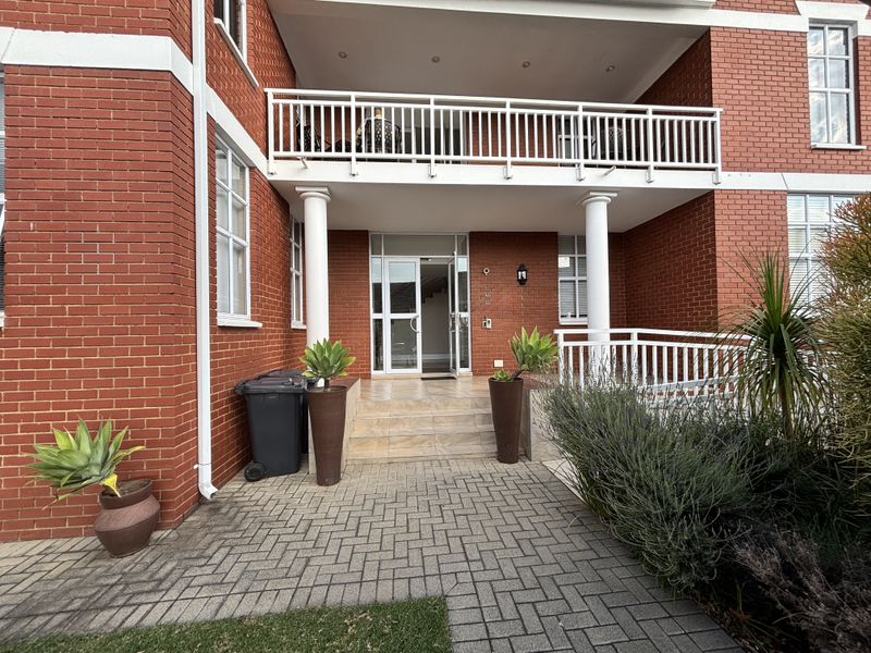Front entrance of the office building featuring a red brick facade, covered balcony with white railings, and a paved approach.