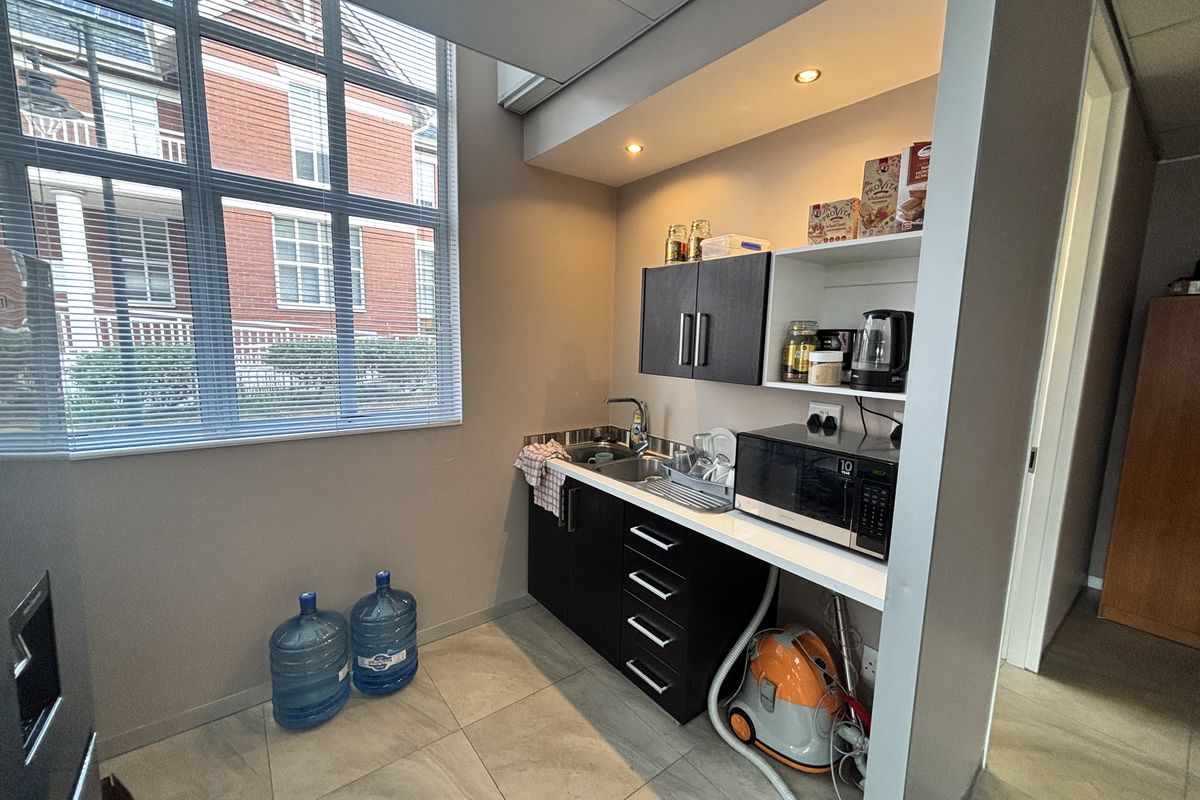 A compact office kitchenette featuring dark cabinetry, a stainless steel sink, recessed lighting, and large windows providing natural light.