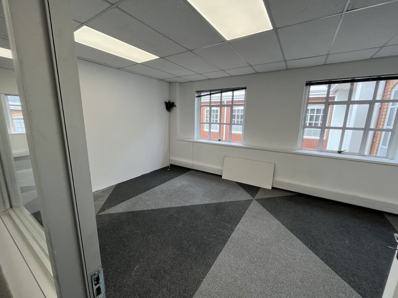 A bright, empty office space featuring patterned carpet tiles, a suspended ceiling with recessed lighting, and large windows providing natural light.