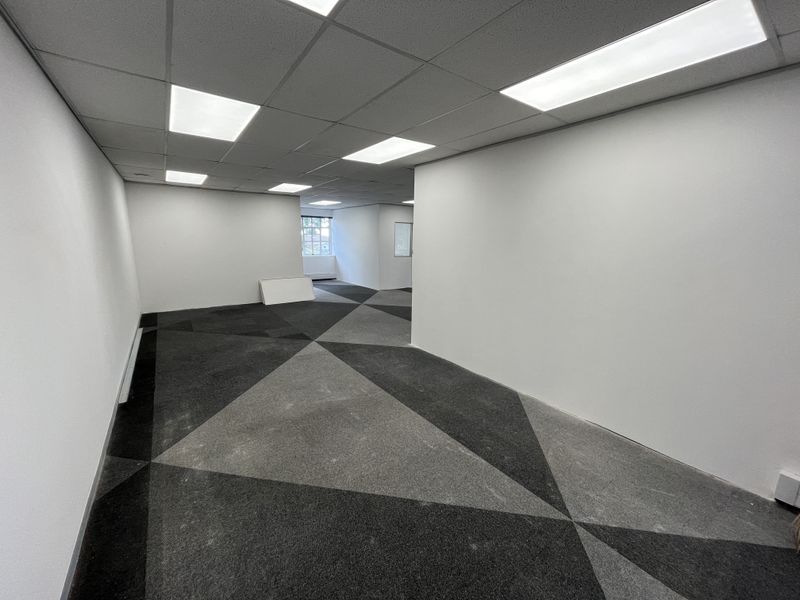 A spacious, vacant office area featuring a suspended ceiling with recessed lighting and modern geometric patterned carpet tiles.