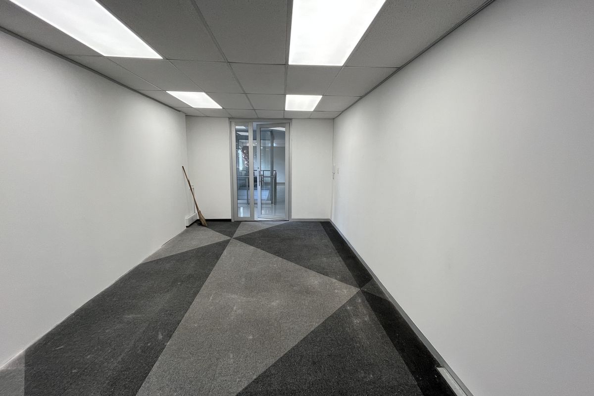 A clean, empty office space featuring a suspended ceiling with recessed lighting, patterned carpet tiles, and a glass panel entrance door.