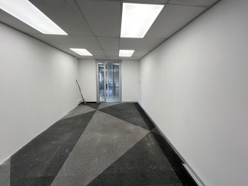 A clean, empty office space featuring a suspended ceiling with recessed lighting, patterned carpet tiles, and a glass panel entrance door.