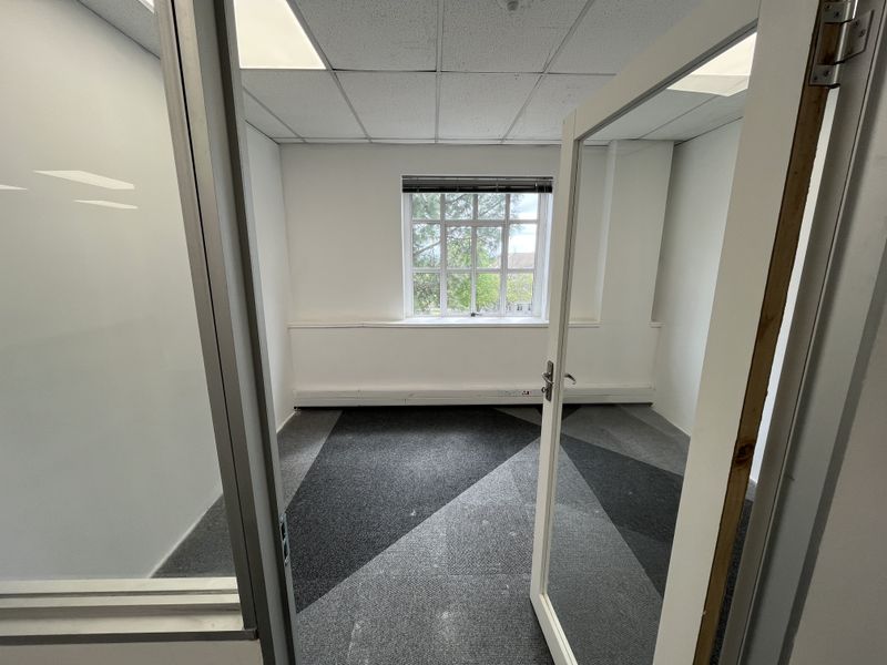 A vacant office space featuring a suspended ceiling, recessed lighting, patterned carpet tiles, and an aluminium-framed glass entrance door.