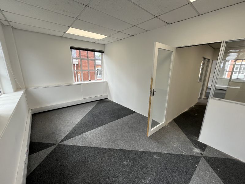 An empty office space featuring a suspended ceiling, recessed lighting, patterned carpet tiles, and a glass partition wall.