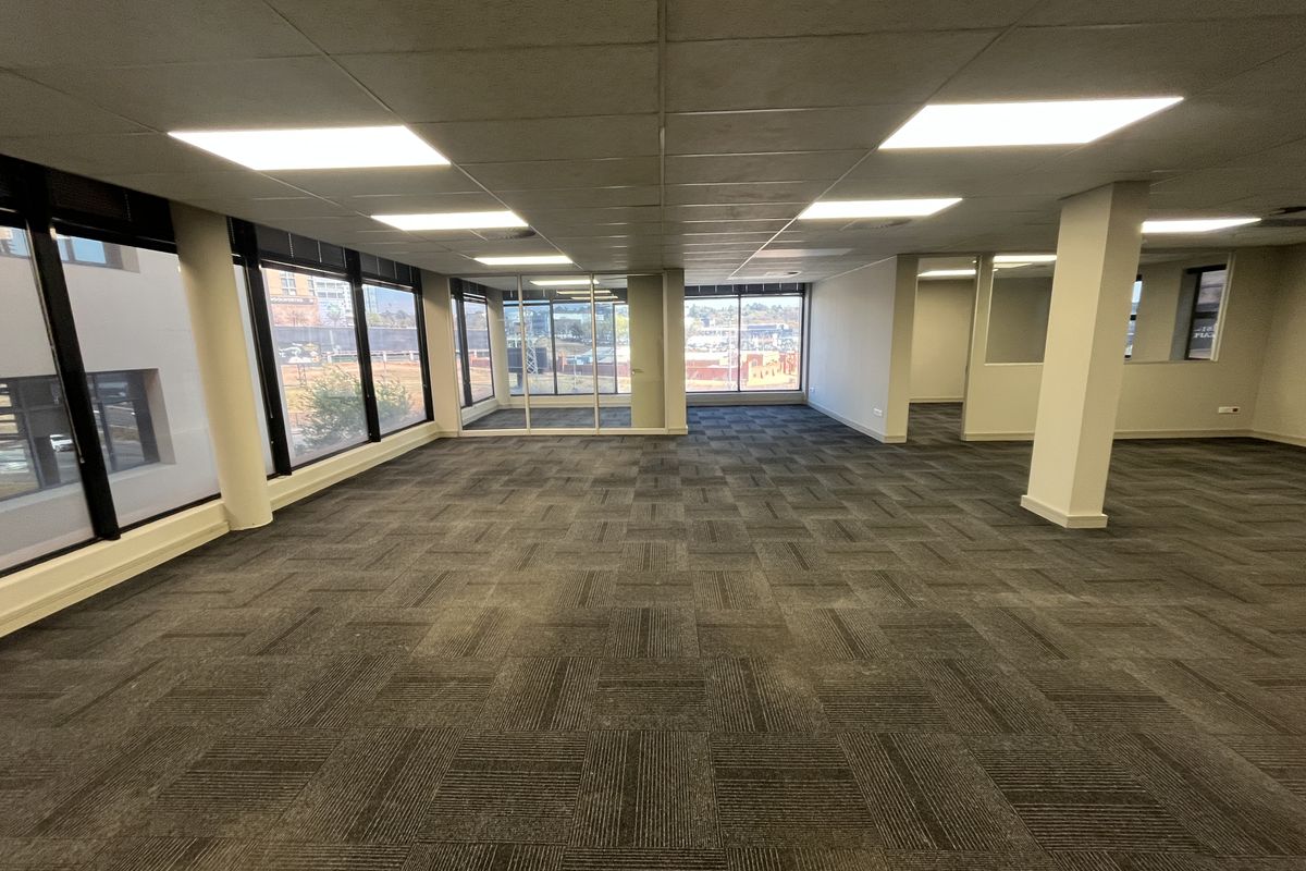 Spacious open-plan office area featuring large windows, carpet tiles, and a suspended ceiling with recessed lighting, offering a bright and professional workspace.