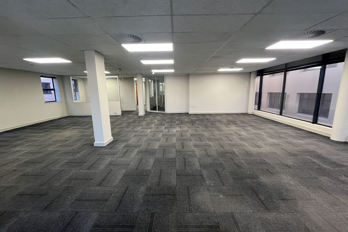 A spacious, open-plan office area featuring neutral carpet tiles, a suspended ceiling with recessed lighting, and large windows providing ample natural light.