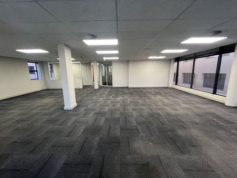 A spacious, open-plan office area featuring neutral carpet tiles, a suspended ceiling with recessed lighting, and large windows providing ample natural light.