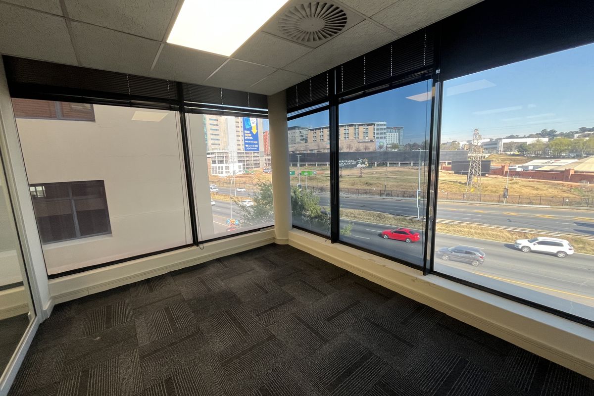 A bright, empty corner office space featuring carpet tiles, suspended ceilings with recessed lighting, and expansive windows offering an elevated urban view.