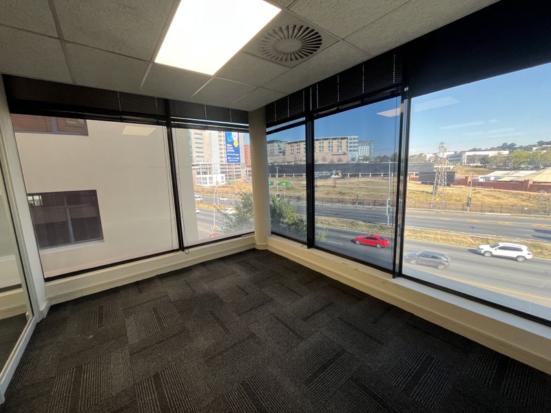 A bright, empty corner office space featuring carpet tiles, suspended ceilings with recessed lighting, and expansive windows offering an elevated urban view.