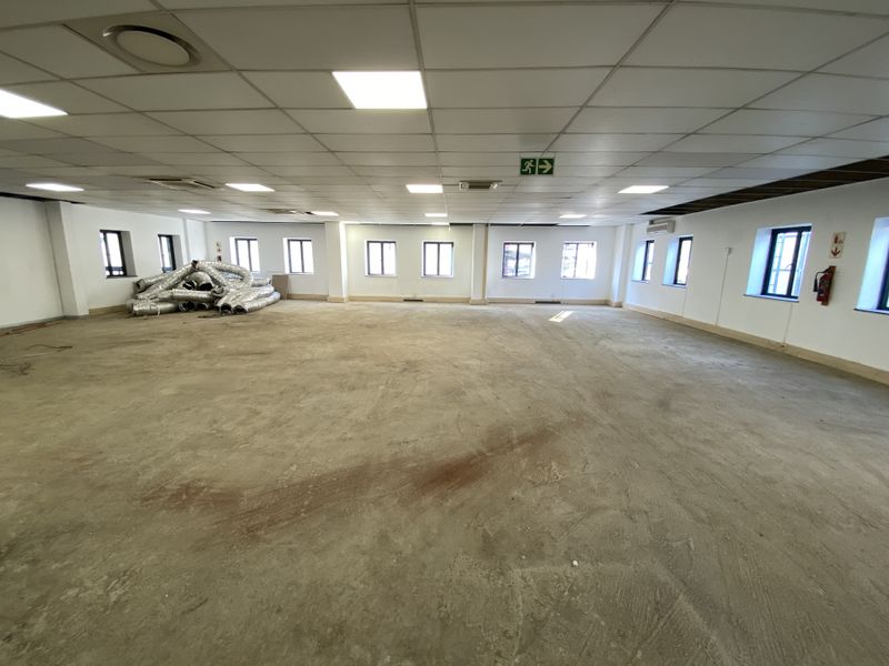 A large, open-plan commercial space featuring a suspended ceiling, recessed lighting, and multiple windows, currently awaiting final flooring installation.