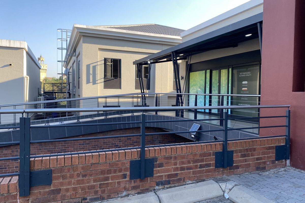 Exterior view of a retail storefront featuring a modern metal pedestrian bridge, stainless steel balustrades, and a brick retaining wall.