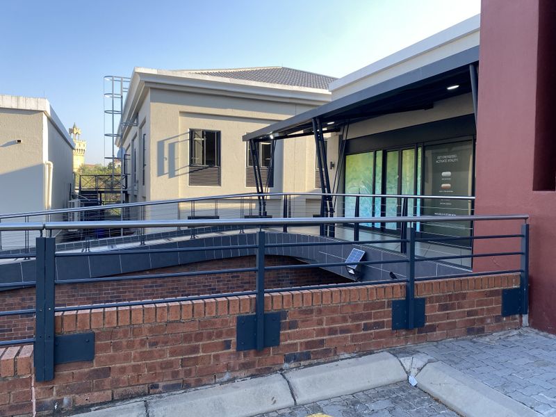 Exterior view of a retail storefront featuring a modern metal pedestrian bridge, stainless steel balustrades, and a brick retaining wall.