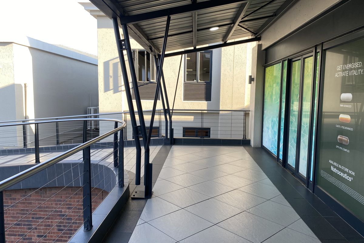A modern covered balcony area featuring tiled flooring, a metal roof structure, and stainless steel railings, providing access to the retail space.