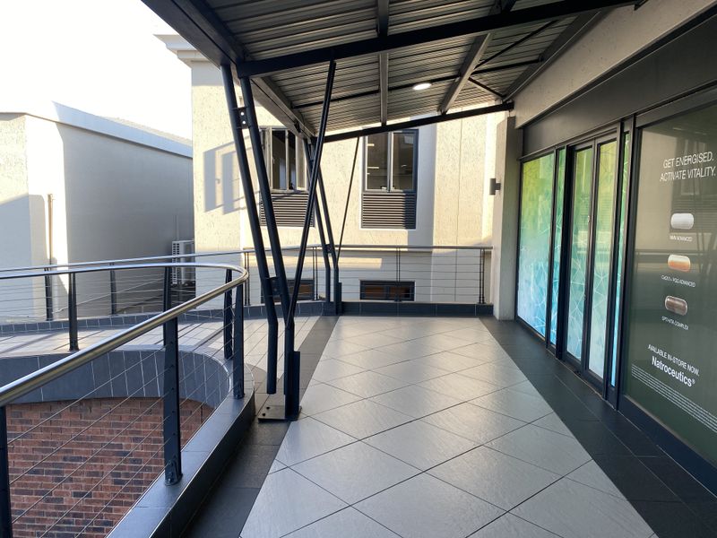 A modern covered balcony area featuring tiled flooring, a metal roof structure, and stainless steel railings, providing access to the retail space.