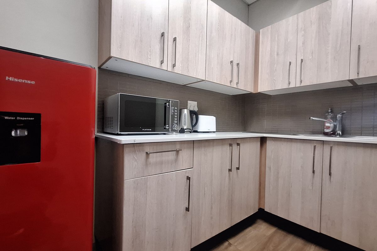 A compact office kitchenette featuring light wood-grain cabinetry, a stainless steel sink, and a tiled backsplash.
