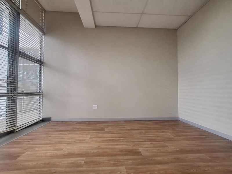 A bright, empty office space featuring modern laminate wood flooring, large windows with venetian blinds, and a clean, neutral interior finish.