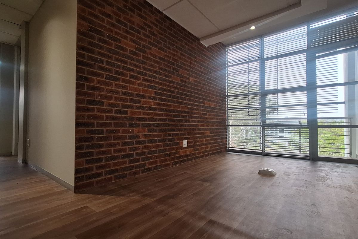 A bright, empty office space featuring an exposed brick wall, large floor-to-ceiling windows with blinds, and modern laminate flooring.