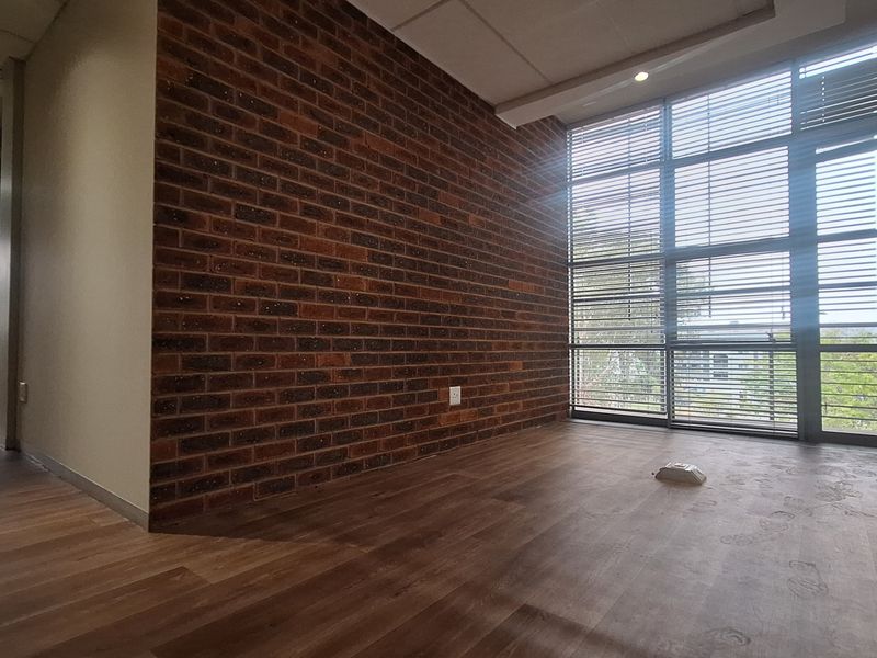 A bright, empty office space featuring an exposed brick wall, large floor-to-ceiling windows with blinds, and modern laminate flooring.