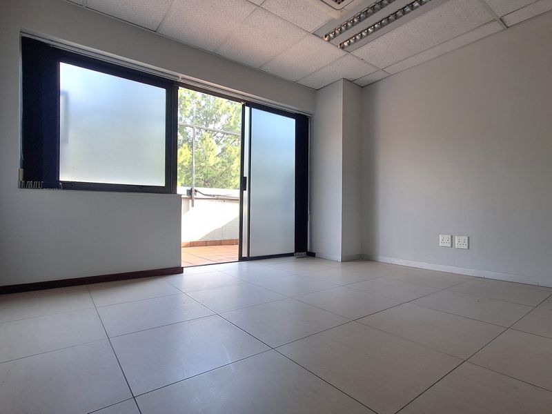 A bright, empty office space featuring tiled flooring, a suspended ceiling with integrated lighting, and a sliding glass door leading to a balcony.