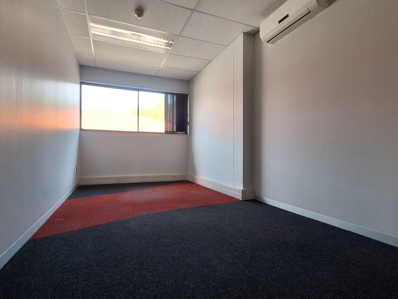 A vacant office space featuring a suspended ceiling, recessed lighting, a wall-mounted air conditioning unit, and dual-tone carpet flooring.