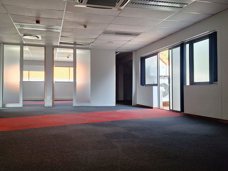 An empty open-plan office space featuring suspended ceilings, glass partitions, and carpeted flooring with access to an outdoor area.