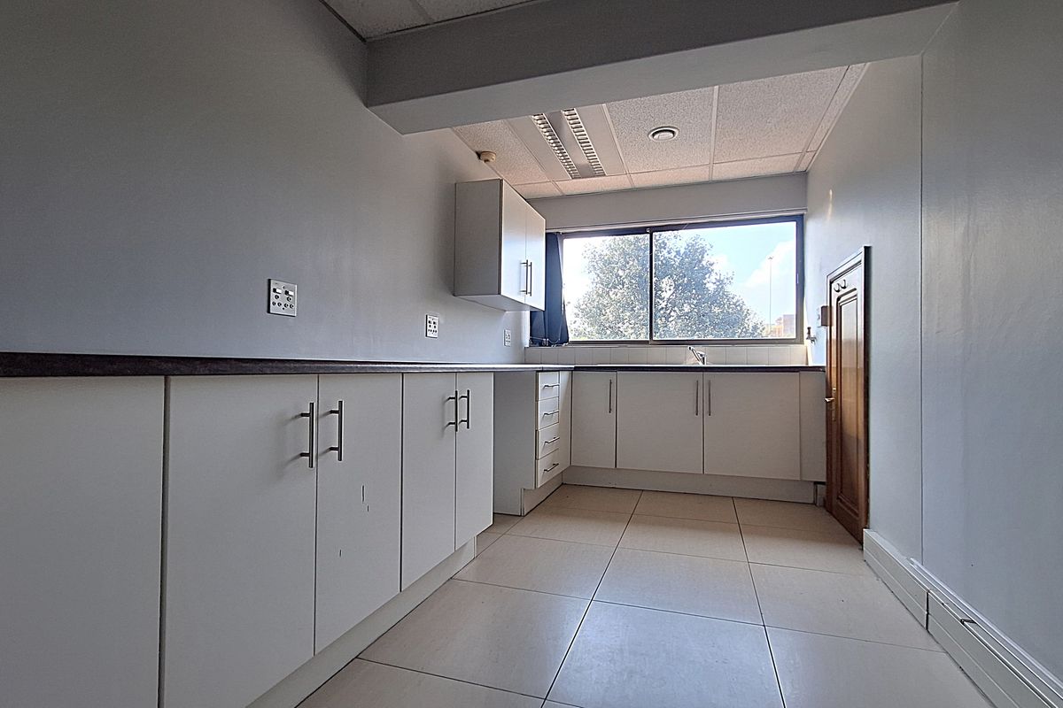 A functional, empty office kitchen featuring white built-in cabinetry, laminate countertops, tiled flooring, and a suspended ceiling with integrated lighting.