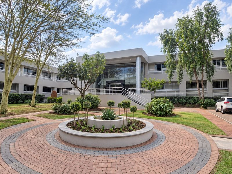 A professional exterior view of a modern office building featuring a glass entrance, paved circular driveway, and well-maintained landscaped gardens.