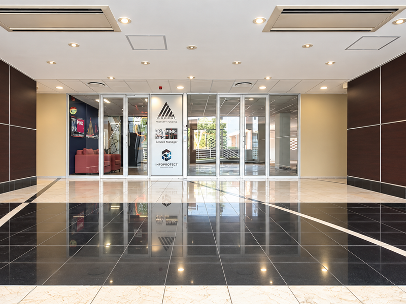 A professional office building entrance lobby featuring polished tiled flooring, dark wood wall paneling, and glass entry doors with integrated ceiling lighting.
