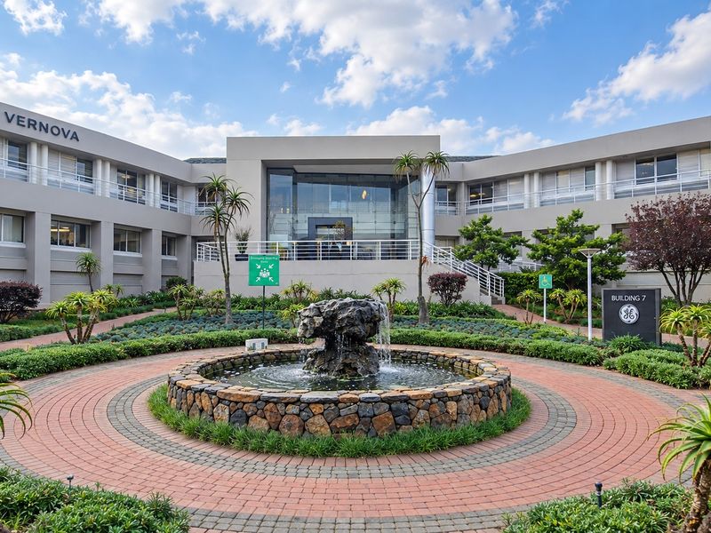 Professional exterior view of a modern office building featuring a central stone fountain, landscaped gardens, and a prominent glass entrance facade.