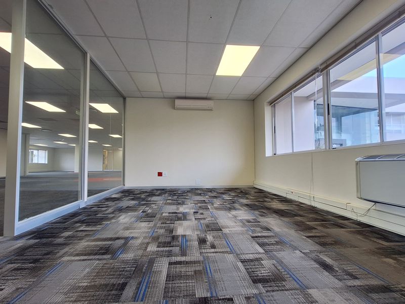 A bright, vacant office space featuring carpet tiles, a glass partition wall, suspended ceilings with recessed lighting, and integrated air conditioning units.