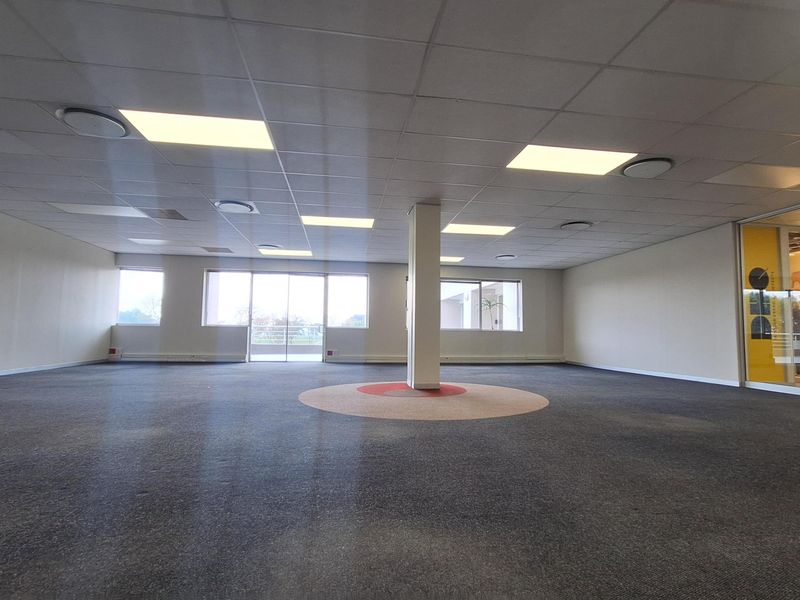 A spacious, open-plan office area featuring a suspended ceiling, recessed lighting, and a central support column with carpeted flooring throughout.
