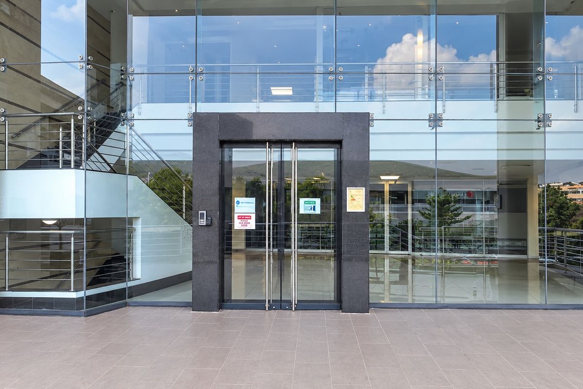 Modern office building entrance featuring a glass curtain wall facade, stone-clad doorway, and tiled exterior landing.