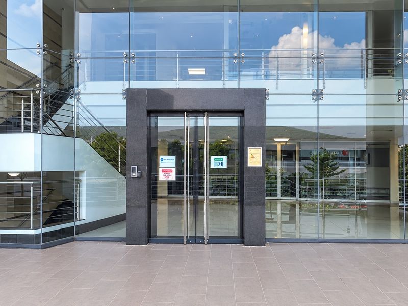 Modern office building entrance featuring a glass curtain wall facade, stone-clad doorway, and tiled exterior landing.