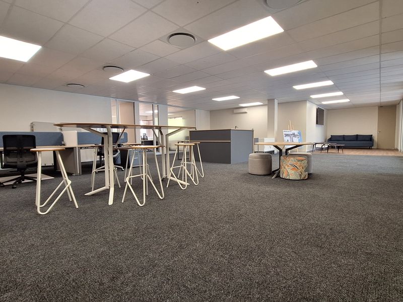 A spacious, open-plan office area featuring a suspended ceiling with recessed lighting, carpeted flooring, and modern glass partitions.
