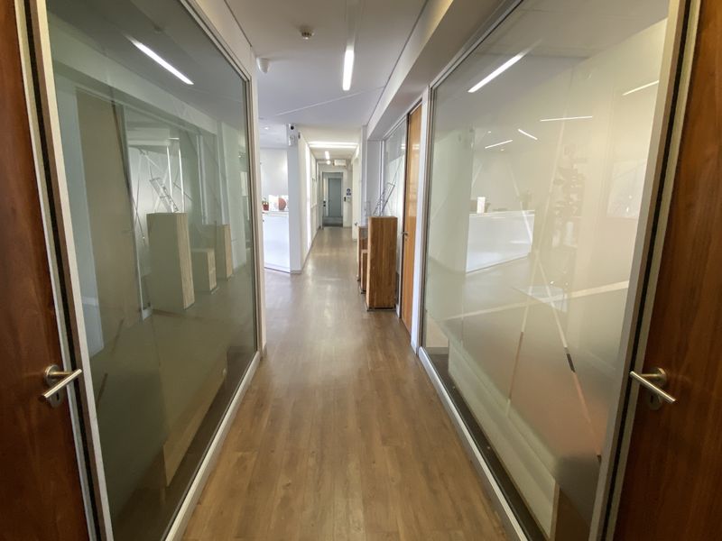 A modern office hallway featuring wood veneer doors, laminate flooring, and glass partition walls with frosted detailing, leading to an open-plan workspace.