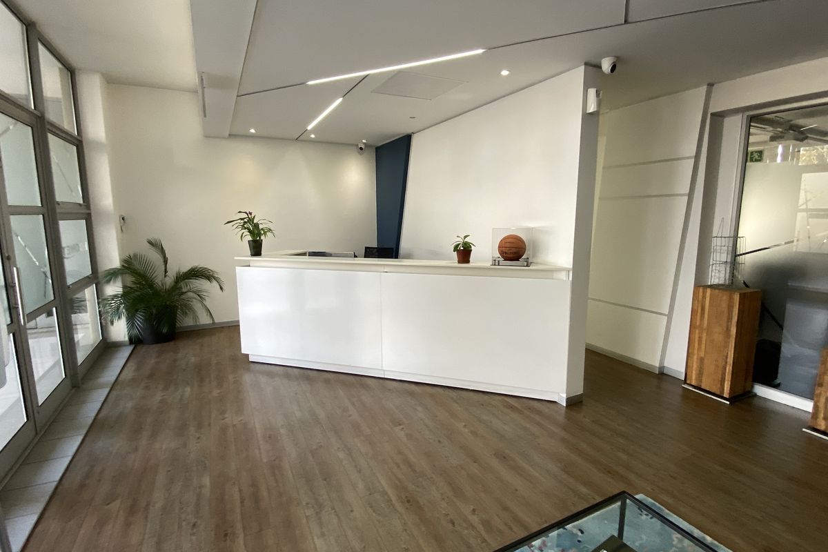 Modern office reception area featuring a clean white desk, wood-look laminate flooring, and large glass-paneled entrance doors.