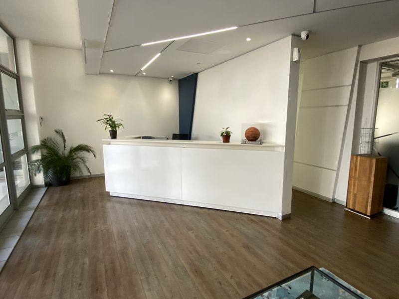 Modern office reception area featuring a clean white desk, wood-look laminate flooring, and large glass-paneled entrance doors.