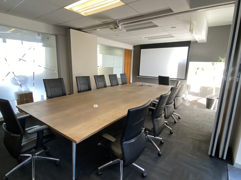 A professional boardroom featuring a large meeting table, integrated projector screen, glass partitioning, and modern carpet tiles in a well-lit office environment.