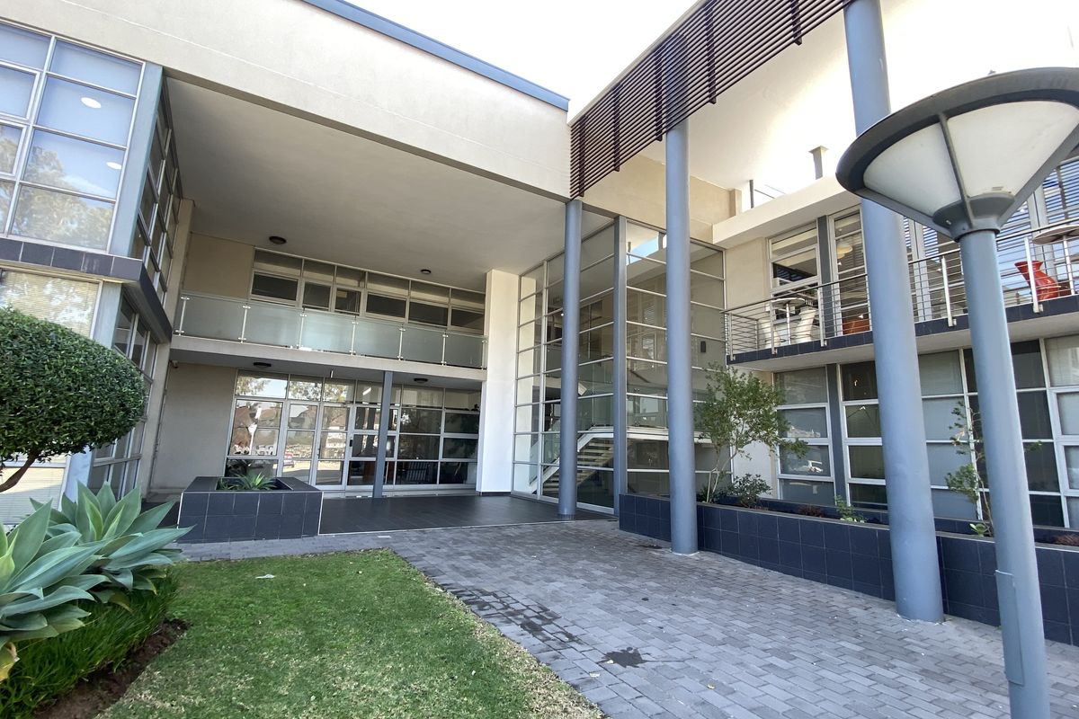 Modern office building entrance featuring a glass facade, steel structural columns, paved walkway, and integrated balcony areas with glass balustrades.