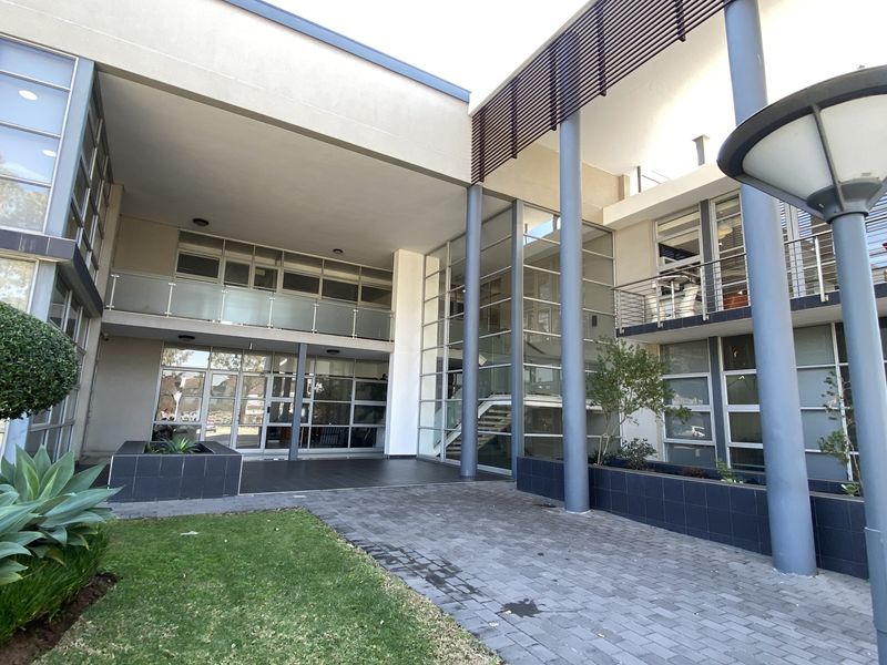 Modern office building entrance featuring a glass facade, steel structural columns, paved walkway, and integrated balcony areas with glass balustrades.