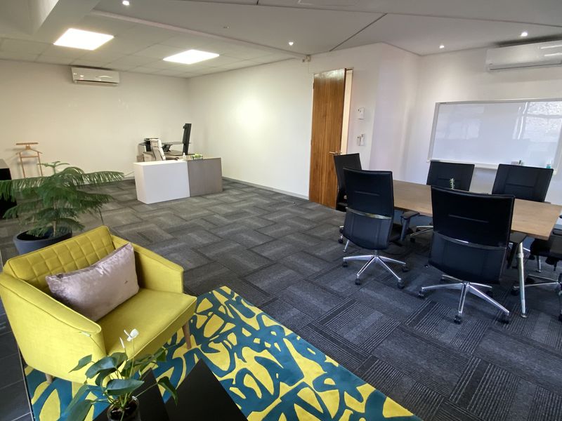 A spacious office area featuring carpeted flooring, recessed ceiling lights, a wall-mounted whiteboard, and a split-system air conditioning unit.