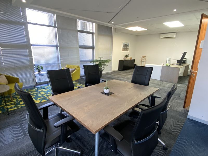 A well-lit professional office space featuring large windows with blinds, carpeted flooring, and integrated air conditioning, suitable for meetings or individual work.