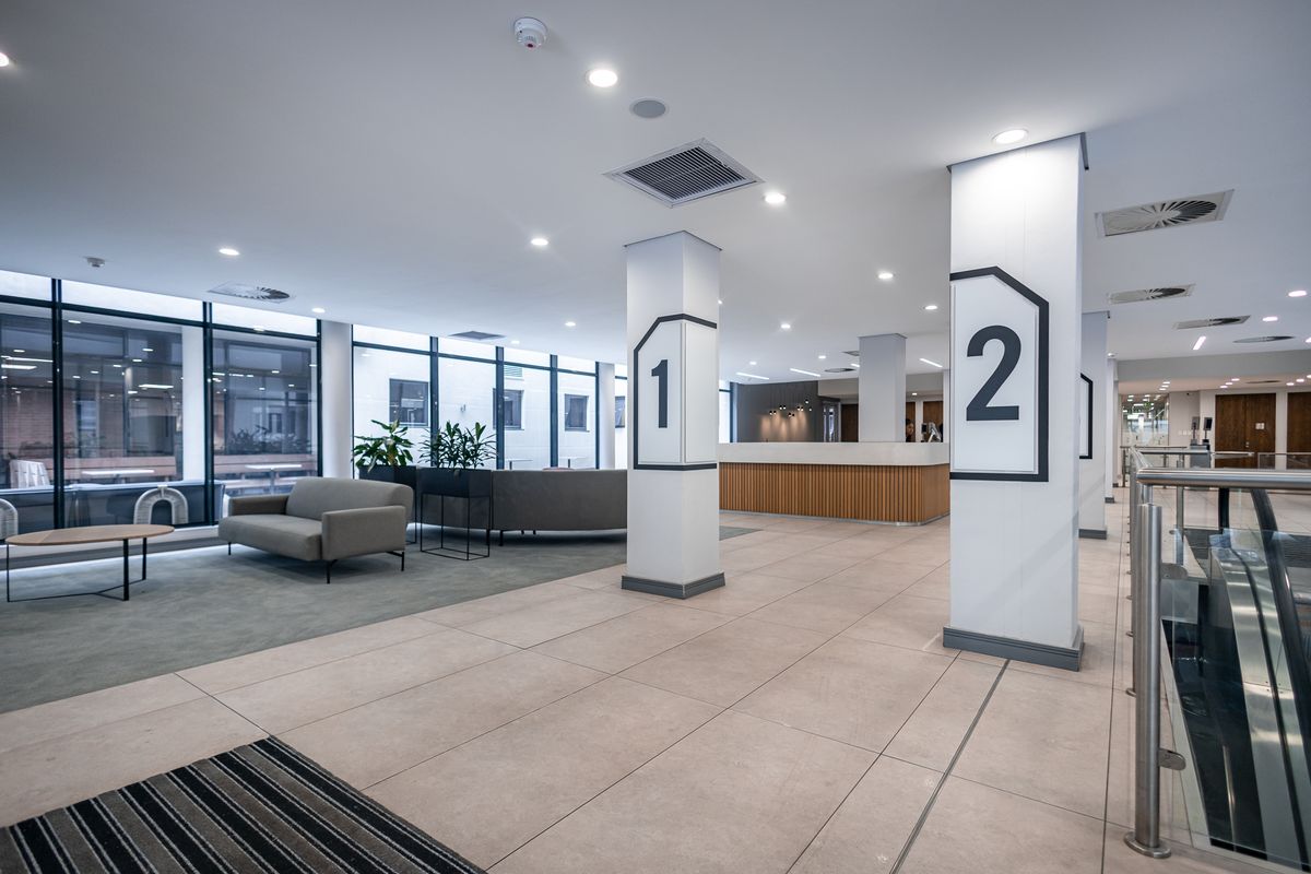 Modern office lobby featuring tiled flooring, structural columns with signage, recessed lighting, and a professional reception area with floor-to-ceiling windows.