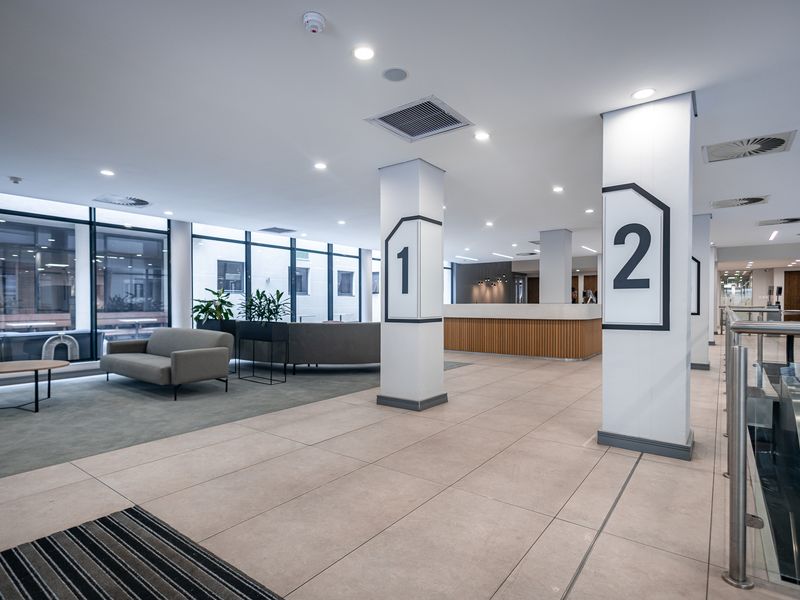Modern office lobby featuring tiled flooring, structural columns with signage, recessed lighting, and a professional reception area with floor-to-ceiling windows.
