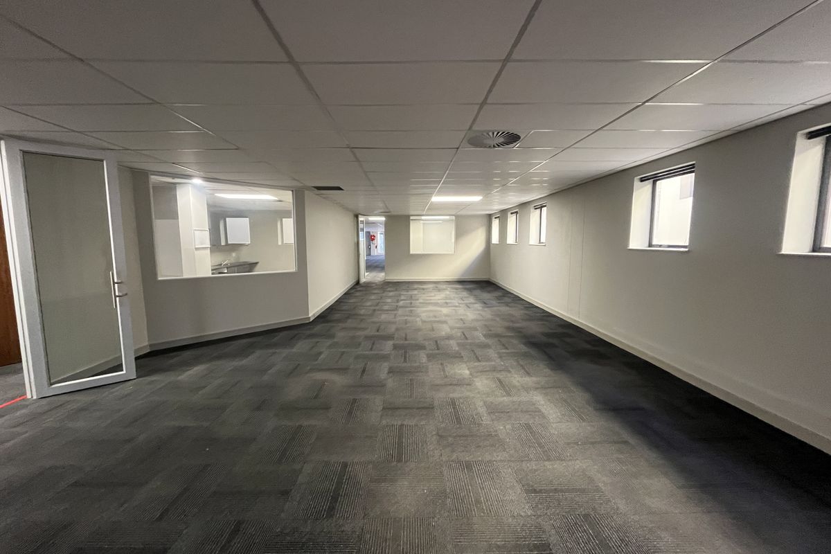 A spacious, empty office area featuring neutral carpet tiles, a suspended ceiling with recessed lighting, and a glass partition wall.
