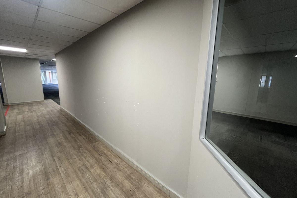 A vacant office hallway featuring wood-look laminate flooring, a suspended ceiling with recessed lighting, and a glass partition wall.