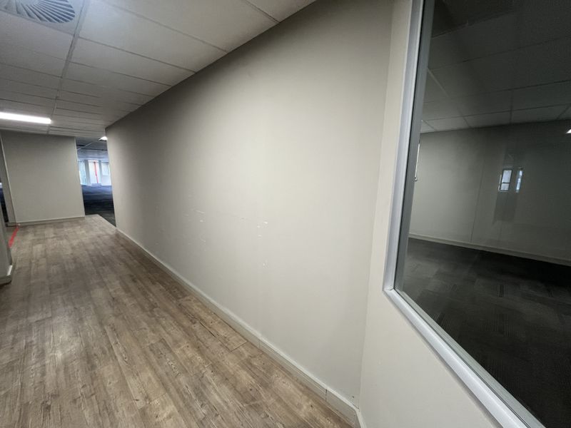 A vacant office hallway featuring wood-look laminate flooring, a suspended ceiling with recessed lighting, and a glass partition wall.
