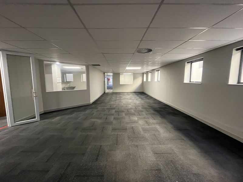 A spacious, empty office area featuring neutral carpet tiles, a suspended ceiling with recessed lighting, and a glass partition wall.