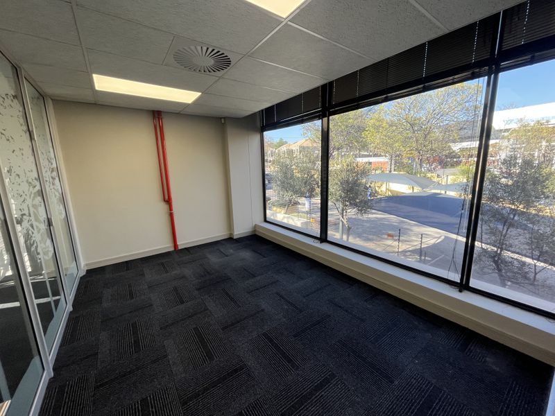 A bright, vacant office space featuring large windows, carpet tiles, a suspended ceiling, and a glass partition wall, offering a professional work environment.
